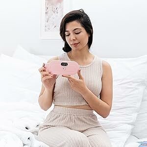 A woman holding a pink Portable Heating pad while sitting on the bed.