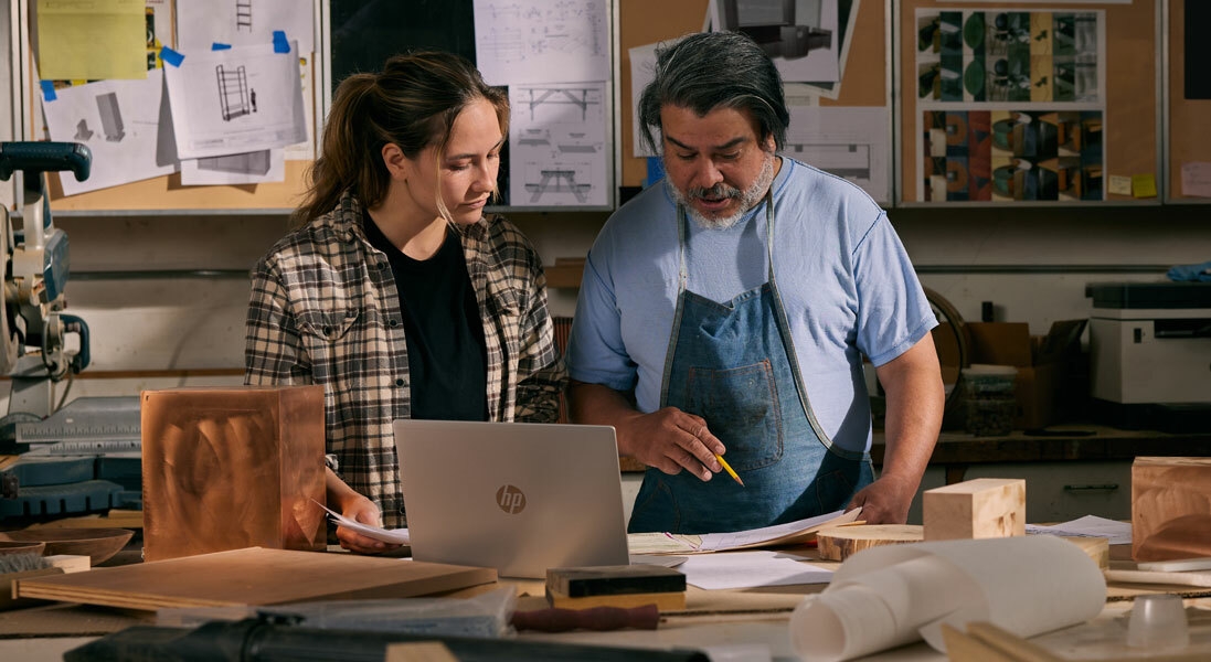 A man and a women are engaged in an important discussion while working on HP Laptop surrounded by wooden materials and drawings.