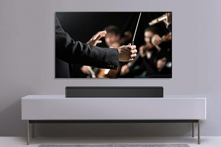 A TV is shown on a gray wall and LG Soundbar below it on a gray shelf. TV shows a conductor conducting an orchestra.